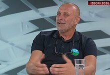 Човек говори на ТВ емисији уз стаклени чаши и намештај у позадини.