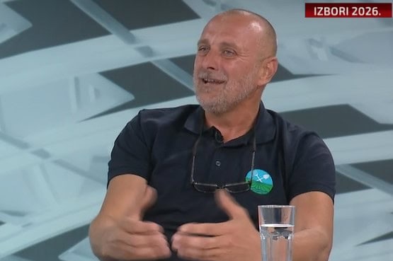 Човек говори на ТВ емисији уз стаклени чаши и намештај у позадини.