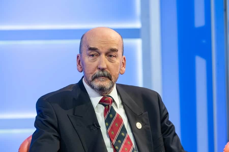 Političar u tamnom odelu sa crvenim prugastim kravatom sedi na panel diskusiji.