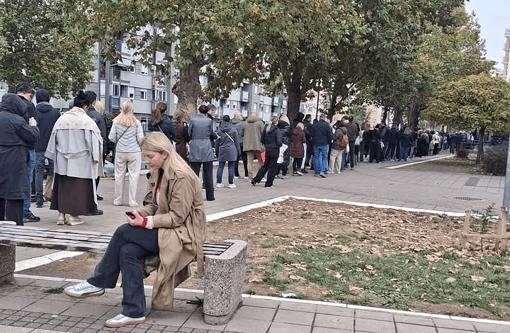 Линија људи чека испред изборне фирме; женa седа на клупи у предворју.