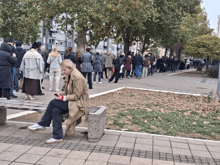 Линија људи чека испред изборне фирме; женa седа на клупи у предворју.