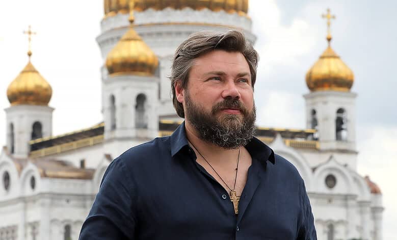 Портрет који приказује мушкарца са брежуљастом брадом испред православне цркве са златним куполама.