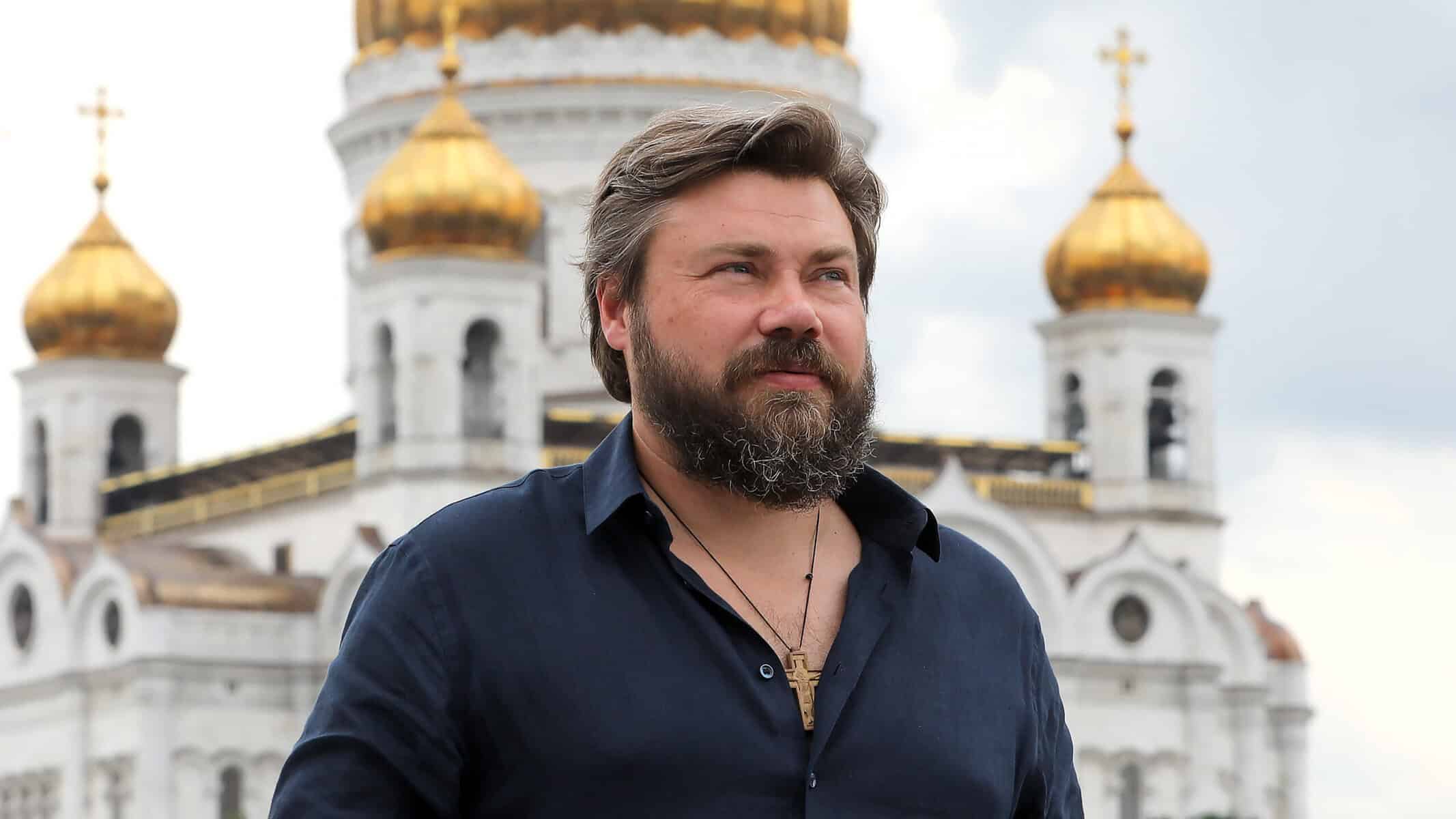 Портрет који приказује мушкарца са брежуљастом брадом испред православне цркве са златним куполама.