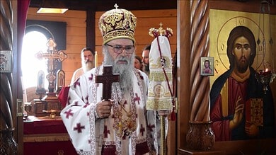 Високи црквени службеник у цркви са крстом и иконама у позадини.